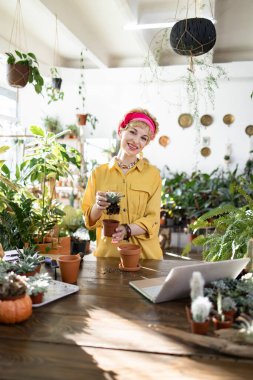 Gülümseyen bir kadın, yemyeşil bir yer ve ahşap bir masadaki dizüstü bilgisayarla çevrili sulu bir sıvıyı temsil eder.