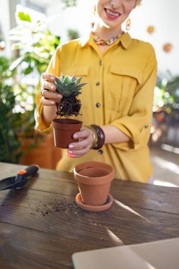 Gülümseyen bir kadın, küçük bir sulu tencereyi dikkatlice yeni bir tencereye nakleder. Bahçıvanlık sürecinin tadını çıkarır.