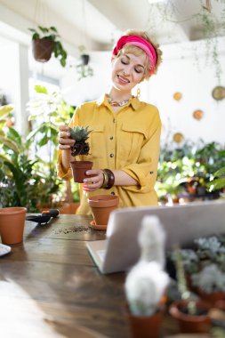 A smiling woman is shown transplanting a succulent into a new pot, surrounded by lush greenery