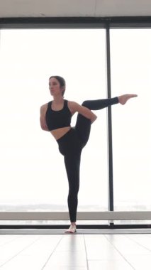 Siyah giysili bir kadın büyük bir pencerenin yanında zarif ve esnek bir şekilde dikey yoga pozu veriyor.