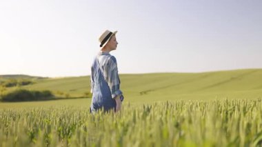 Hasır şapka ve kot pantolon giyen genç bir kadın açık bir gökyüzünün altında geniş bir buğday tarlasında duruyor.