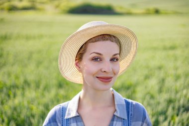 Hasır şapka ve ekose gömlek giyen gülümseyen bir kadın güneşin altında yemyeşil bir tarlada duruyor.