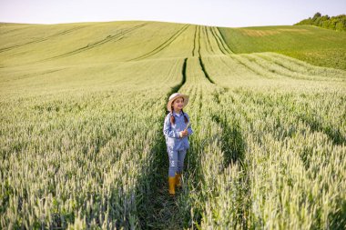 Hasır şapkalı ve sarı çizmeli genç bir kız açık gökyüzünün altındaki geniş yeşil buğday tarlasında duruyor.