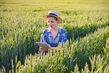 A young woman wearing a straw hat and overalls stands in a lush wheat field, examining data on a tablet computer