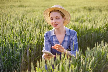 A woman wearing a straw hat and overalls uses her mobile phone while standing in a lush green wheat field during the day