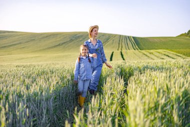 Bir anne ve küçük kızı açık bir gökyüzünün altındaki yemyeşil buğday tarlasında el ele yürüyorlar.