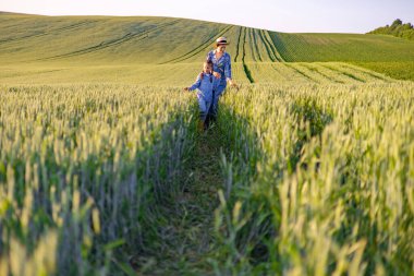 Bir anne ve kızı berrak bir gökyüzünün altındaki yemyeşil buğday tarlasında birlikte güneşli bir günün tadını çıkarıyorlar.
