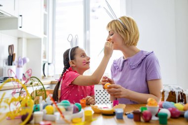 Tavşan kulağı takan bir anne ve kız, Paskalya hazırlıkları sırasında birbirlerini renkli resimlerle boyamakla eğleniyorlar.