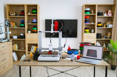 Bir 3D yazıcı aktif olarak masanda iplik makaraları ve 3 boyutlu bir model gösteren bir dizüstü bilgisayarı olan kırmızı bir nesne yaratıyor.