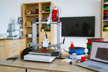 Modern bir 3D yazıcı, ahşap masadaki iplik makaraları ve aletlerle çevrili ısıtılmış yatağında kırmızı bir nesne yaratıyor.