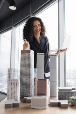 Gülümseyen bir mimar panoramik ofis penceresinin yanında gökdelen modellerini gösterirken başparmağını kaldırır