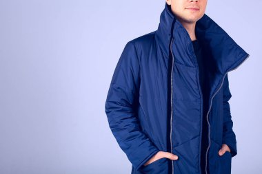 Gri arka plandaki adam mavi ceketli. Elleri kalın bir ceketin cebinde. Sakallı ve sıkı giyinmiş bir adam. Şık, kalın kıyafetler.