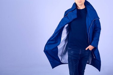 Gri arka plandaki adam mavi ceketli. Elleri kalın bir ceketin cebinde. Sakallı ve sıkı giyinmiş bir adam. Şık, kalın kıyafetler.