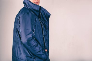 Gri arka plandaki adam mavi ceketli. Elleri kalın bir ceketin cebinde. Sakallı ve sıkı giyinmiş bir adam. Şık, kalın kıyafetler.