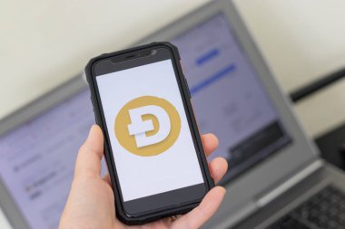 Dogecoin popüler bir kripto para birimidir. iPhone ekranındaki Dogecoin logosunu kapat. Değerinin grafiği olan bir dizüstü bilgisayarın önünde dur..