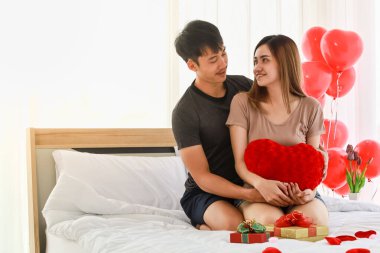 Çift ve Sevgililer Günü Konsepti. Gülümseyen Asyalı adam yatak odasında kız arkadaşına kırmızı kalp yastığı, güzel hediye kutusu ve balonla sarılıyor..