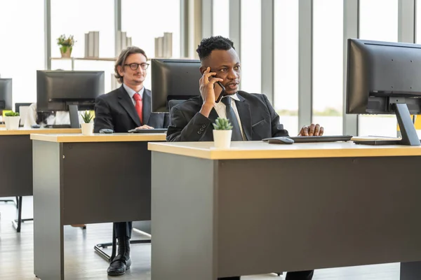 Diversity Business and Work palce concept. African businessman talking on call with mobile smartphone in office with desktop computer.