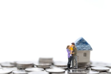 Ev ve Aile Kavramı. Ebeveyn erkek, kadın ve çocuk, beyaz arka planda izole edilmiş mini ev oyuncaklarıyla bozuk para yığınının üzerinde öpüşüp sarılıyorlar..