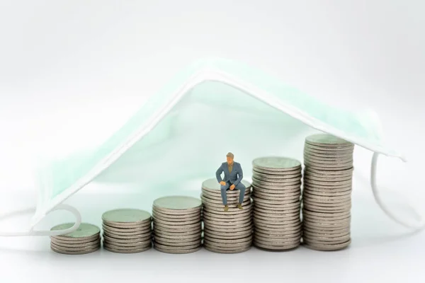 Business, Money, Covid-19 and Healthcare Concept. Businessman miniature figure people sitting on stack of silver coins under surgical face mask on white background.