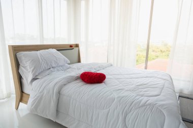 Beyaz perdeli beyaz yatak odası, beyaz yastıklar, kırmızı kalp şeklindeki yastık ve pencere manzaralı yorgan..