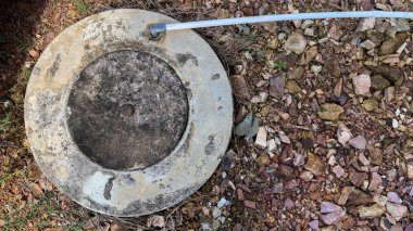 Metal kulplu yıpranmış beton bir kuyu örtüsü çakıl ve kir üzerine yatar, doğal ışıklandırma ve kaba dokular ile kırsal bir açık hava manzarası yaratır inşaat veya açık hava projeleri için mükemmeldir..