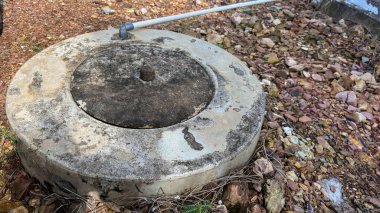 Üzerinde metal bir boru bulunan eskimiş beton bir kuyu, dağınık taşlar ve kuru bitki örtüsüyle kayalık bir açık havaya kurulmuş, tarımsal veya kırsal yaşam tarzına uygun kırsal ve kırsal bir atmosferi taşır..