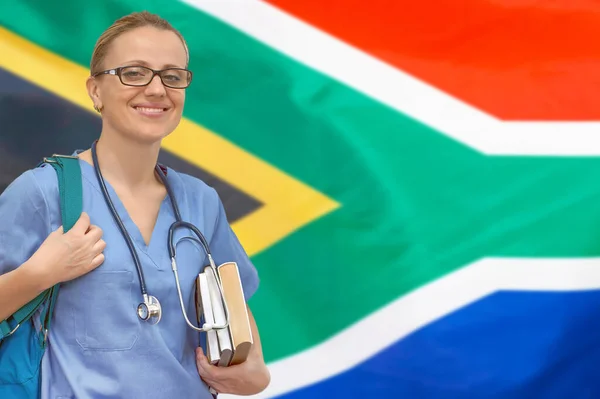 Female student doctor with stethoscope and books in hand on the South Africa flag background. Medical education concept. Medical learning in South Africa