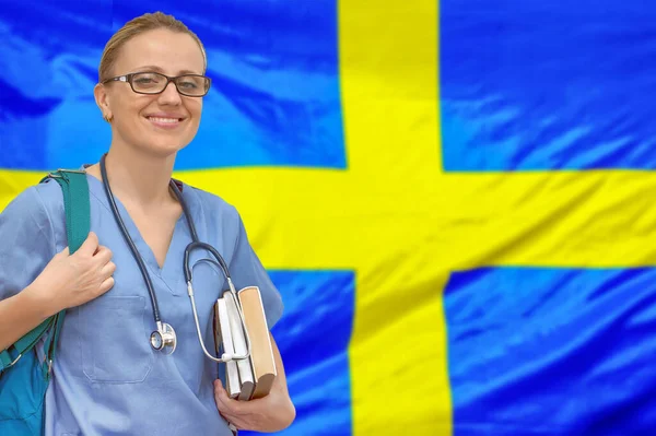 Female student doctor with stethoscope and books in hand on the Sweden flag background. Medical education concept. Medical learning in Sweden