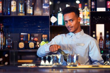 Afrikalı barmen bir şişe viski açar. Arkasında alkol şişeleri olan raflar.                               