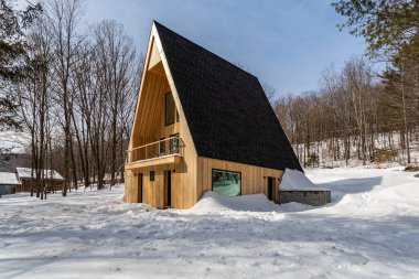 Liberty NY 10 Şubat 2025: Derin kar ve çıplak kış ağaçlarıyla çevrili modern A çerçeveli kulübe, sakin bir kırsal alanda kırsal ahşap mimariyi gösteriyor.