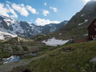 Bremer Hutte 'nin gölü, eriyen kar dilleri ve karla kaplı dağ tepeleri, yemyeşil çayır, mavi gökyüzü arka planı. Bahar sonu güneşli öğleden sonra, canlı renkler.
