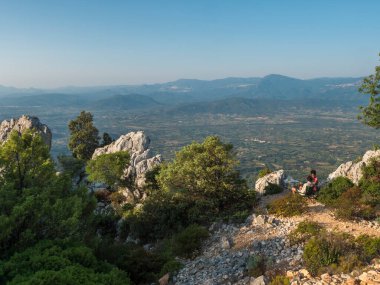 Baunei, Sardinya, İtalya, 17 Eylül 2020: Baunei perspektifinde yeşil ağaçlar, kireçtaşı kayaları, verimli tarlalar ve ovalarla kahvaltı yapan genç çift. Yaz masmavi gökyüzü