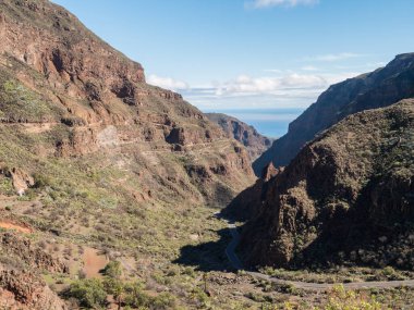 Barranco de Guayadeque 'in sarp duvarları ve okyanusa doğru dönen bir yolu var. Gran Canaria, Kanarya Adası, İspanya. Güneşli bir gün, mavi gökyüzü, beyaz bulutlar...