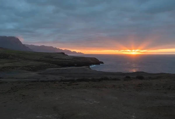 Belo pôr do sol na costa atlântica rochosa na parte noroeste da ilha de Gran Canaria. Sol vermelho alaranjado com raios descendo para o oceano. Nuvens azuis escuras e falésias no fundo. — Fotografia de Stock