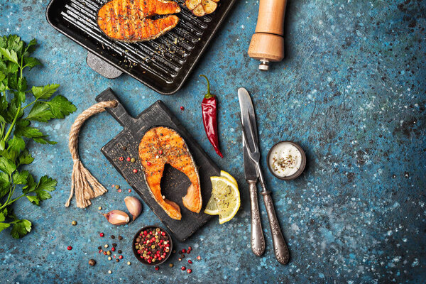 Top view of grilled red fish salmon steak on cast iron skillet or pan with spice, fried garlic, lemon, parsley, pepper mill and tasty sauce on blue concrete background