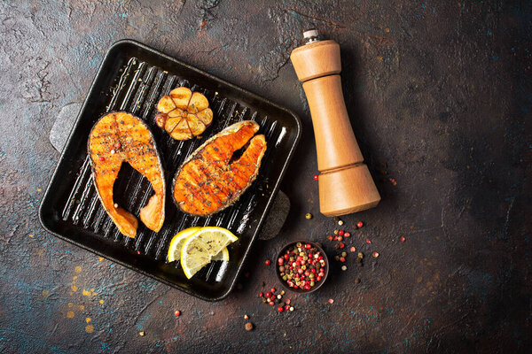 Top view of grilled red fish salmon steak on cast iron skillet or pan with spice, fried garlic, lemon and pepper mill on dark concrete background
