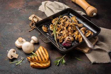 Izgara şampiyon mantarları dökme demir tavada veya tavada baharat, kızarmış soğan, biberiye ve koyu beton arka planda ekmek.
