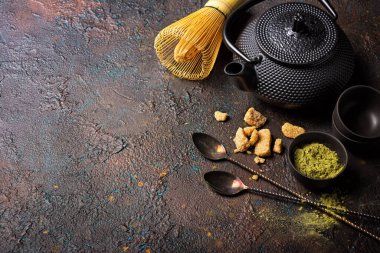 Kâselerdeki organik yeşil kibrit çaydanlığı ya da bambu çırpmalı bardaklar, antika kaşıklar, kamış şekeri ve koyu beton arkaplanda geleneksel siyah Asya çaydanlığı.