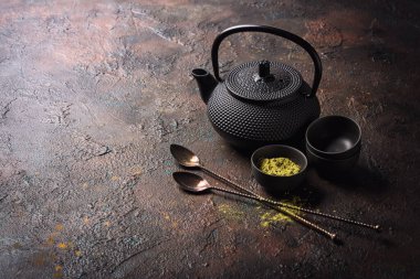 Kâselerdeki organik yeşil kibrit çaydanlığı ya da bambu çırpmalı bardaklar, antika kaşıklar, kamış şekeri ve koyu beton arkaplanda geleneksel siyah Asya çaydanlığı.