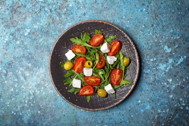 Sağlıklı ve lezzetli vejetaryen yemeklerinin en üst görüntüsü. Taze roka yapraklı salata, kiraz domatesleri, yumuşak peynir ve mavi beton arka planda yeşil zeytinler.