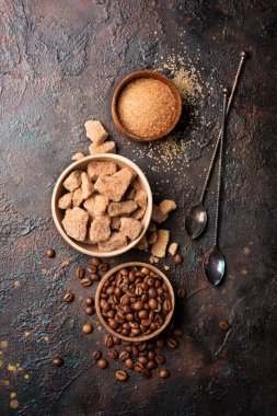 Kahverengi kamış şekeri, yumru ve granüllü kâselerin üst görüntüsü ve kahve çekirdekleri lezzetli içecekler veya koyu beton arka planda içecekler için malzeme olarak kullanılır.