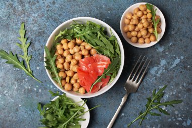 Sağlıklı ve lezzetli yiyecekler. Taze roka yapraklı bir kase pişmiş nohut ve mavi beton arka planda tuzlu kırmızı somon balığı.