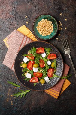 Sağlıklı ve lezzetli vejetaryen yemeklerinin en üst görüntüsü. Taze roka yapraklı salata, kiraz domatesleri, yumuşak peynir, çam fıstığı ve koyu beton arka planda yeşil zeytinler.