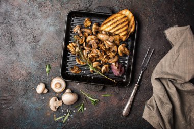 Baharatlı demir tava ya da tavada ızgara şampiyon mantarlarının üst görüntüsü, kızarmış soğan, biberiye ve koyu beton arka planda ekmek.