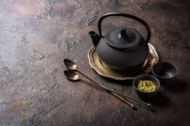 Kâselerdeki organik yeşil kibrit çay tozu veya antika kaşıklı fincanlar ve koyu beton arka planda fotokopi alanı olan geleneksel siyah asyalı çaydanlık.