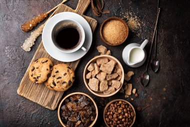 Kahverengi şekerli ve granüllü kâselerin üst görüntüsü, kristal şeker çubukları, kahve çekirdekleri, lezzetli içecekler için süt ve koyu beton arka planda çikolatalı kurabiyeler