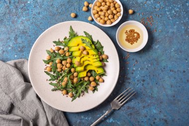 Sağlıklı ve lezzetli vejetaryen yemeklerinin en üst görüntüsü. Pişmiş nohut salatası, taze roka yaprakları ve mavi beton arka planda dilimlenmiş avokado.