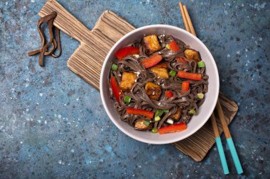 Lezzetli Japon sobalı erişte, kırmızı dolma biber, yeşil soğan, kızarmış tofu peyniri ve mavi beton arka planda susam.