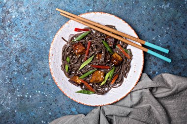 Lezzetli Japon sobalı erişte, kırmızı dolma biber, yeşil soğan, kızarmış tofu peyniri ve mavi beton arka planda susam.