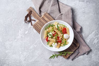 Çeri domatesli lezzetli vejetaryen makarna, yumuşak peynirli mozzarella ve gri arka planda yeşil roka yaprakları.